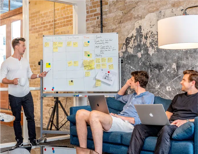 A person presenting a content plan on a whiteboard to two colleagues sat on a sofa with laptops