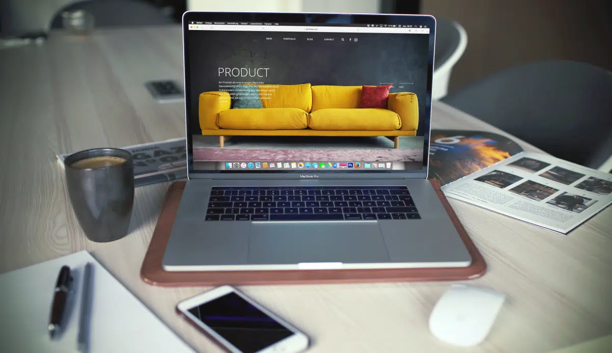 Laptop on a desk in a living room showing a furniture product page with a yellow sofa