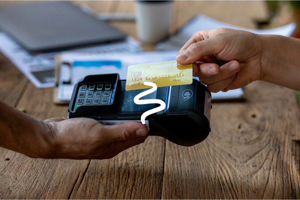 A small business owner taking a card payment from a customer at a wooden table, laptop and notes in the background