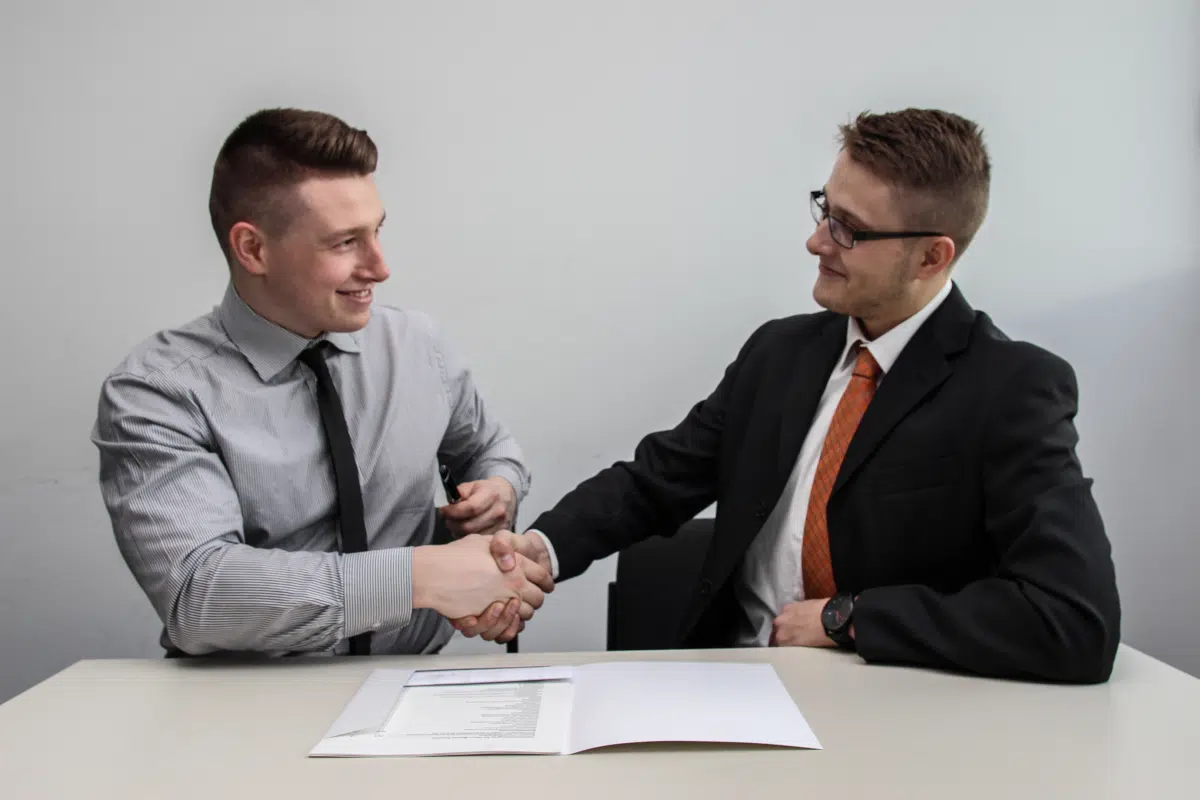 Two men in business attire shaking hands over a desk with an open folder of documents
