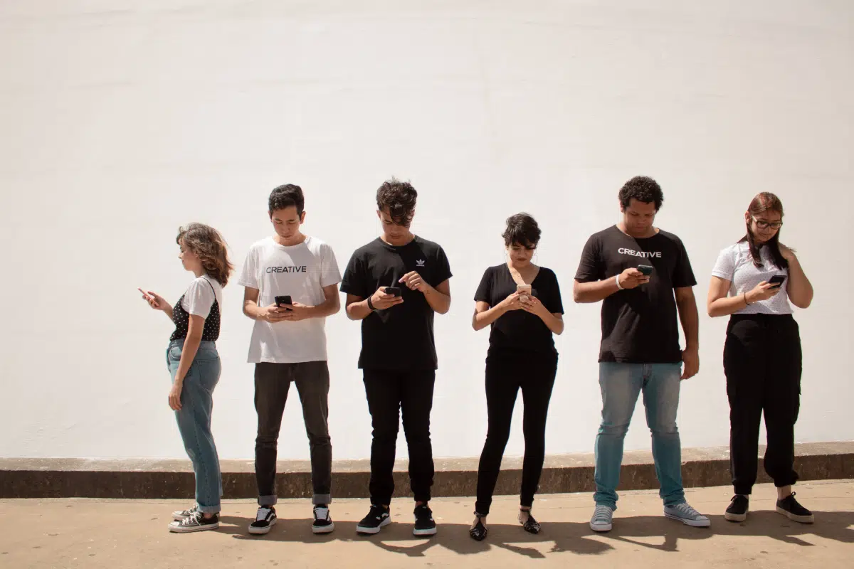 Six people standing against a white wall, all looking down at their phones